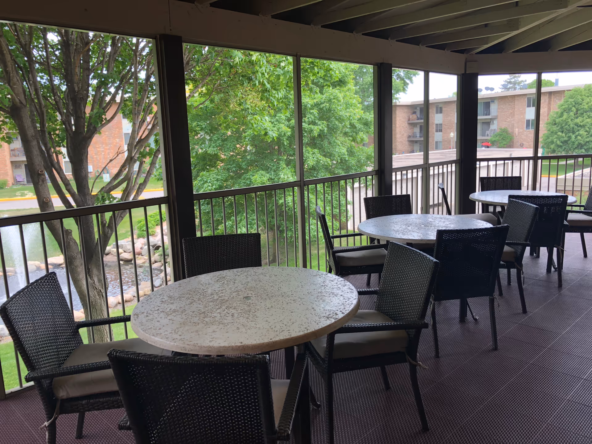 Covered outdoor patio area with round tables and black wicker chairs with cushions, overlooking a landscaped area with trees, a pond with rocks, and brick apartment buildings in the background.