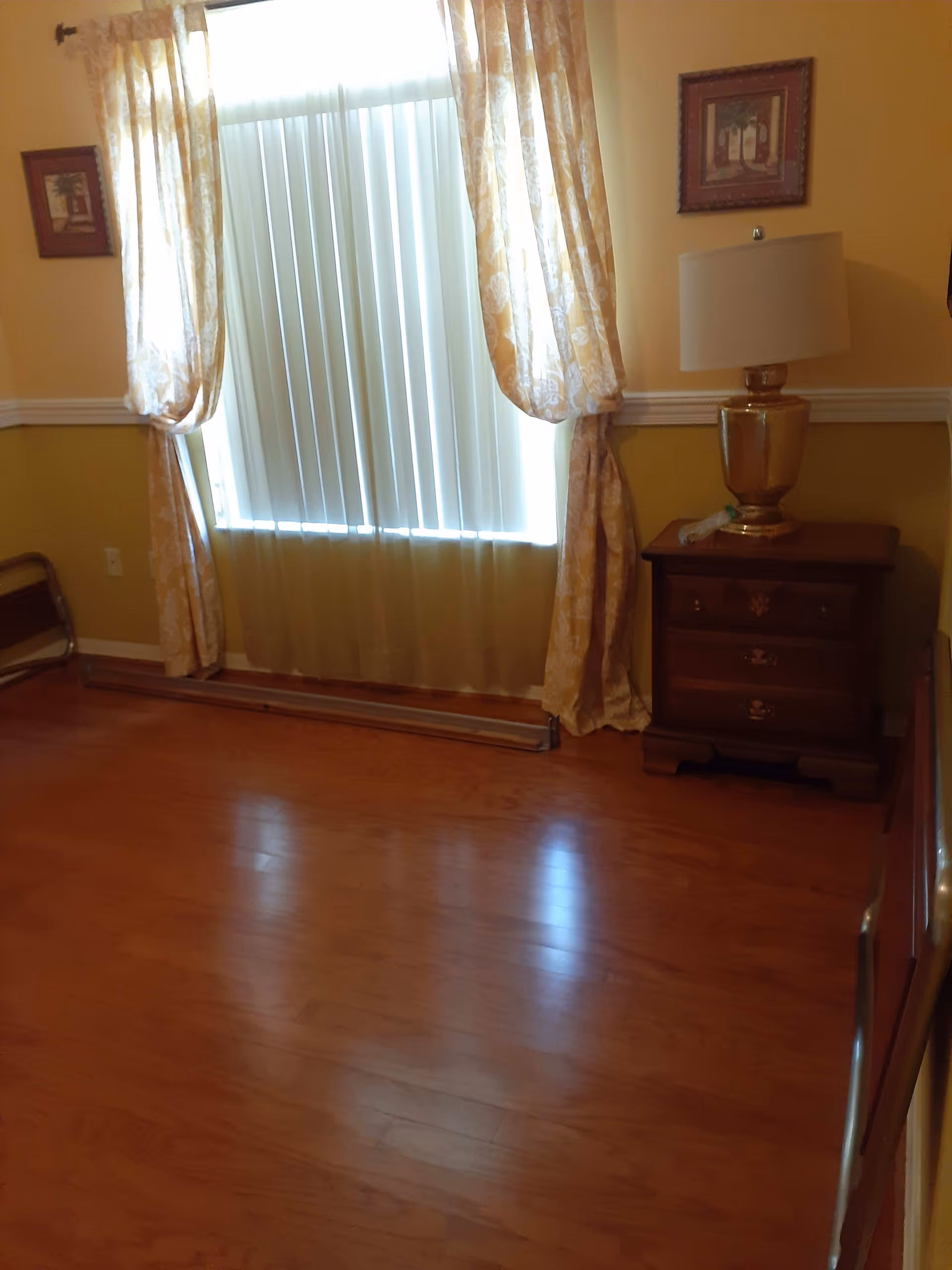 Empty room with hardwood floor, a window with vertical blinds and patterned curtains, and a nightstand topped by a lamp.