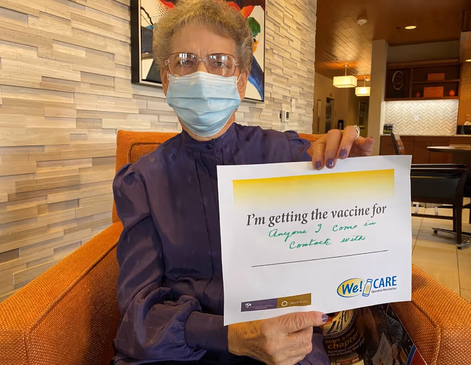 An elderly woman wearing glasses and a face mask sits in an orange armchair inside a facility. She is holding a sign that reads, 'I'm getting the vaccine for Anyone I come in contact with.' The background shows a modern interior with wooden and tiled walls and some furniture.