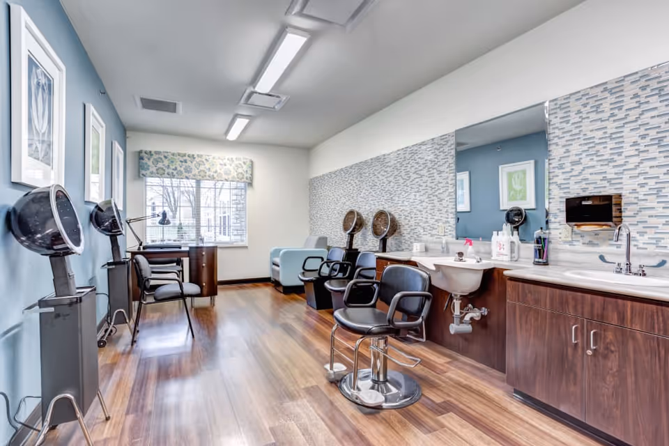 Bright salon-style hair and grooming room with styling chairs, hooded dryers, sinks, and a large mirror.