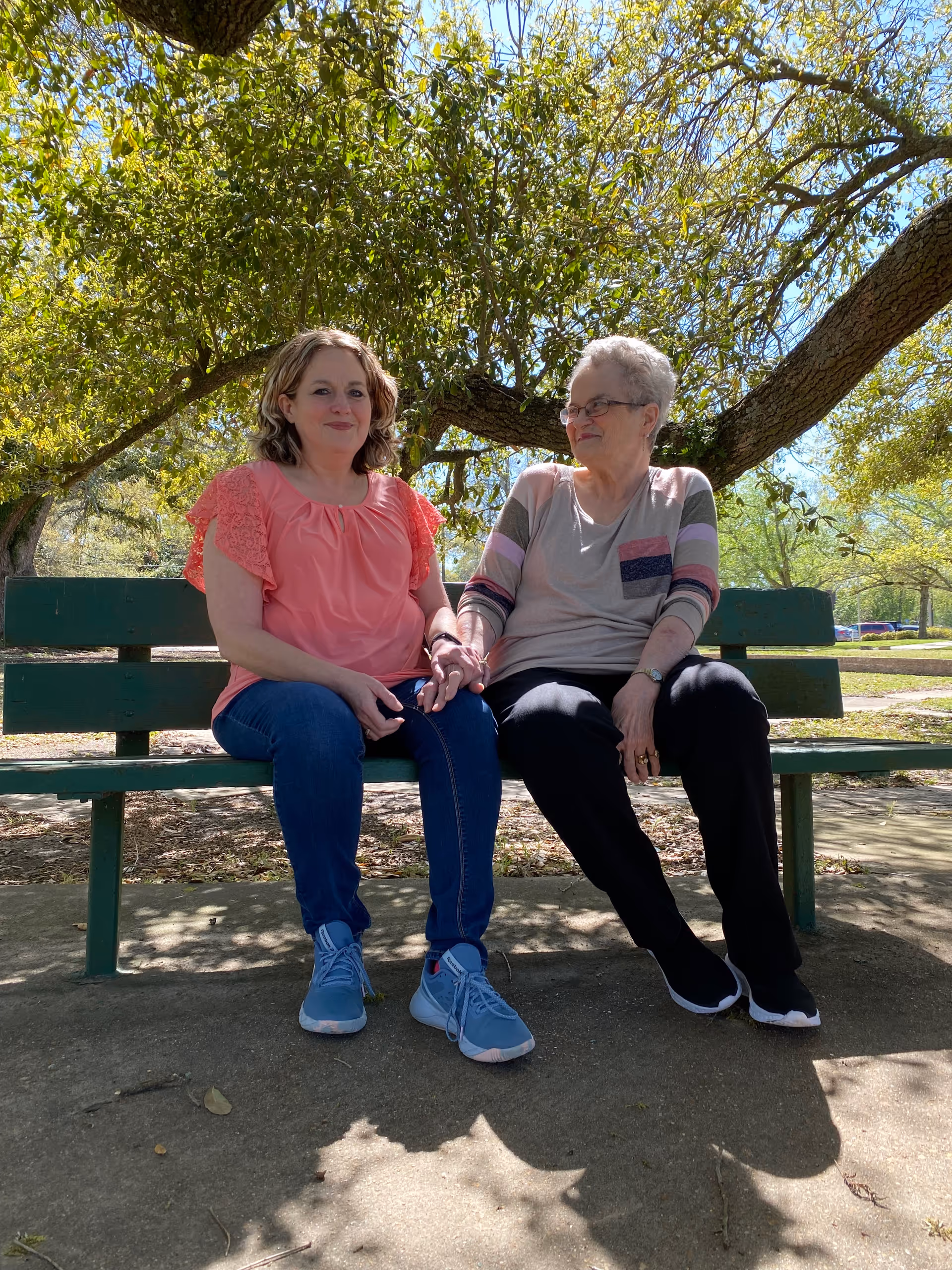 Two women sitting on a green bench outdoors under a tree. One woman is younger, wearing a coral pink blouse and blue jeans, and the other is older, wearing a light gray top with colorful stripes on the sleeves and black pants. They are holding hands and smiling at each other with a sunny park setting in the background.