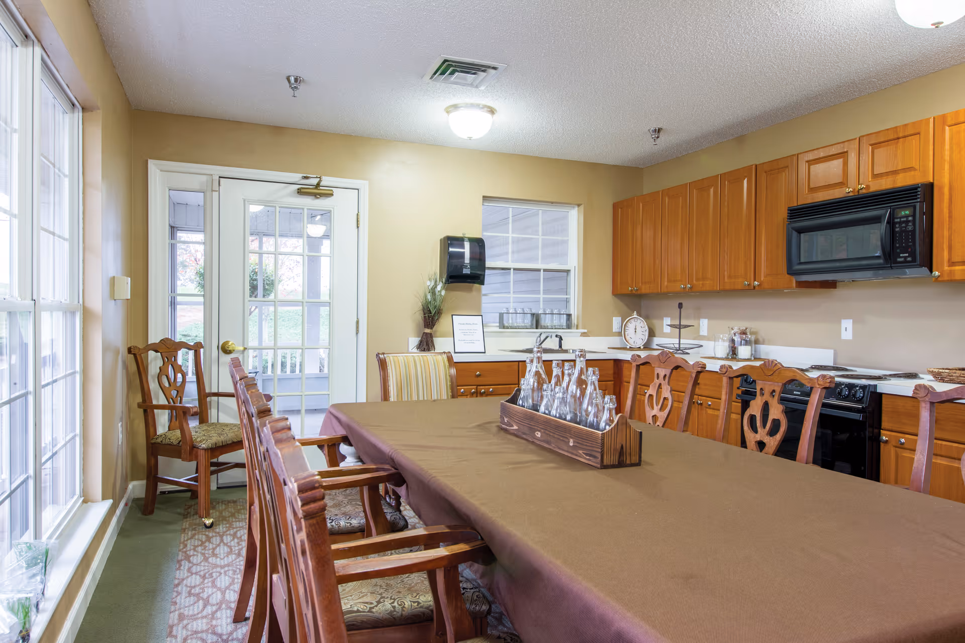 A dining area with a long table covered with a brown tablecloth surrounded by wooden chairs with patterned cushions. The room has large windows on the left side and a door with glass panels at the back. The kitchen area is visible with wooden cabinets, a black microwave, and a stove. A wooden tray with glass bottles is placed on the table.