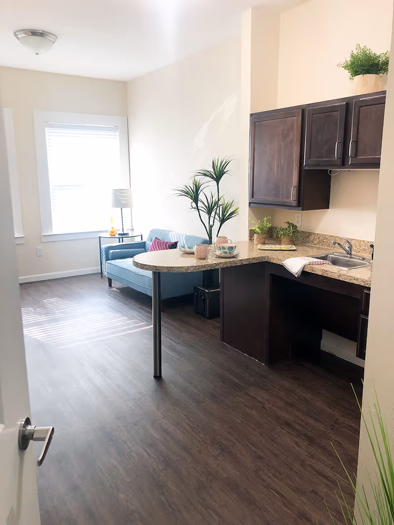 Small open-plan apartment interior with a kitchenette and countertop peninsula next to a blue loveseat by a bright window.