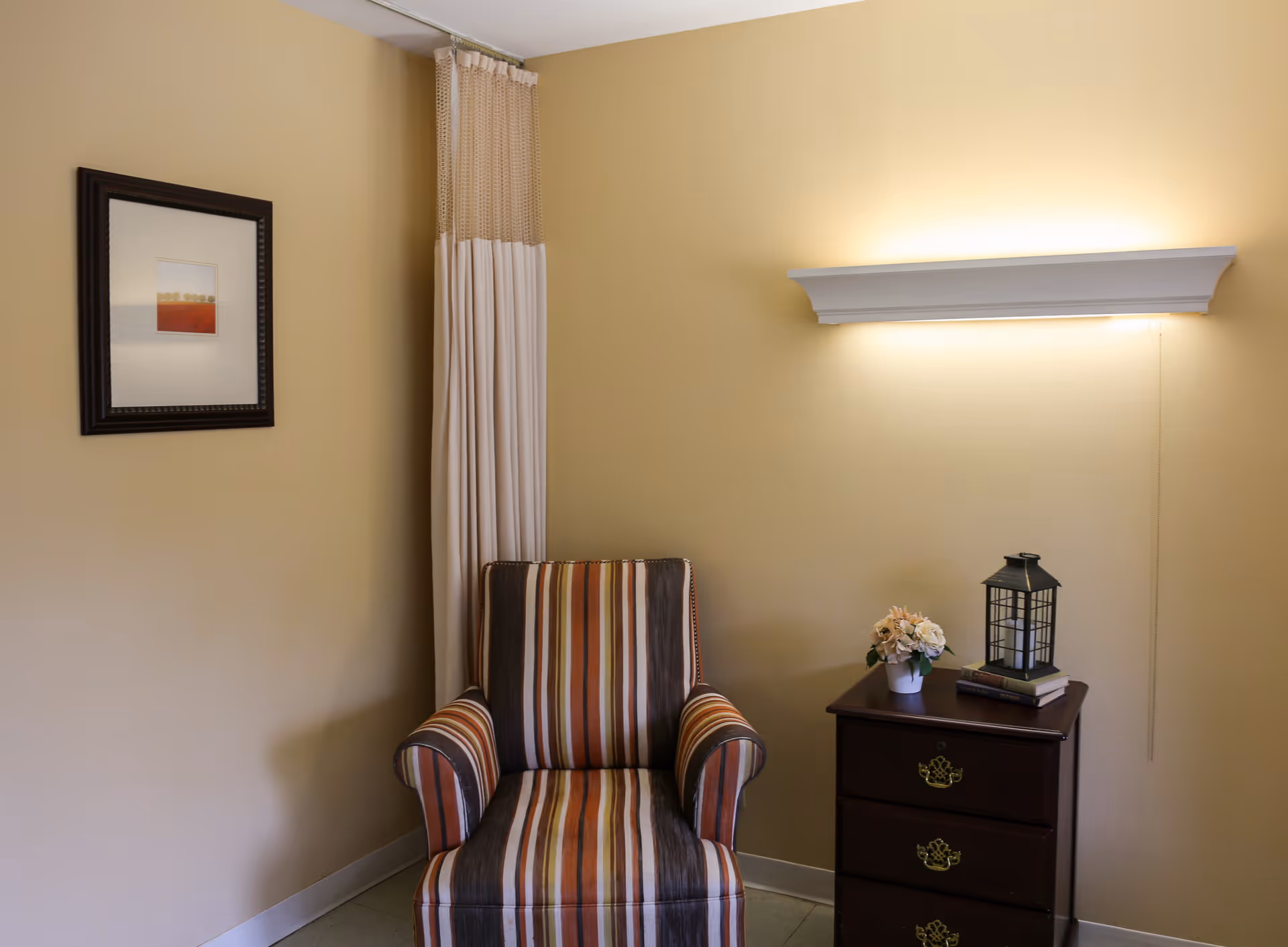 A cozy corner of a room with a striped armchair next to a dark wooden nightstand. On the nightstand, there is a small potted flower arrangement, a decorative lantern, and two stacked books. The wall behind is painted light yellow and features a framed abstract artwork and a wall-mounted light fixture above the nightstand.