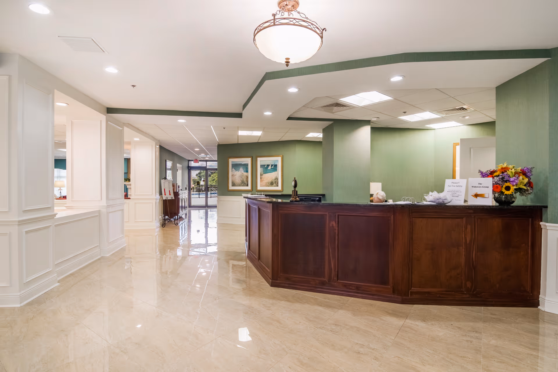 Reception area of Sun Towers Retirement Community featuring a wooden front desk with a flower arrangement and informational signs on top. The walls are painted green with white wainscoting, and the floor is polished beige tile. Two framed pictures hang on the wall behind the desk, and a glass door is visible in the background.