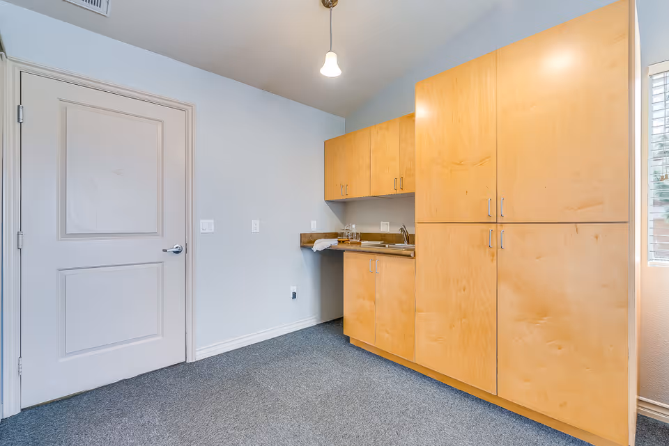 A small kitchenette area with light wood cabinets, a countertop with a sink, and a towel and glassware on the counter. There is a closed white door on the left and a window with blinds on the right, with grey carpeted flooring and a single ceiling light fixture.
