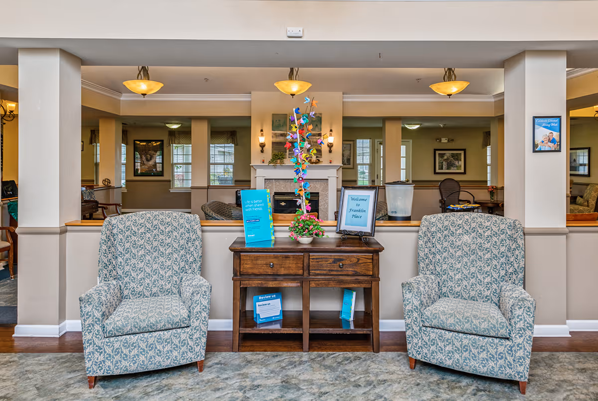 A cozy seating area in a senior living facility with two patterned armchairs flanking a wooden table. The table holds a colorful decorative tree, informational brochures, and a framed sign that reads 'Welcome to Franklin Place.' The background shows a fireplace and additional seating in a warmly lit room.