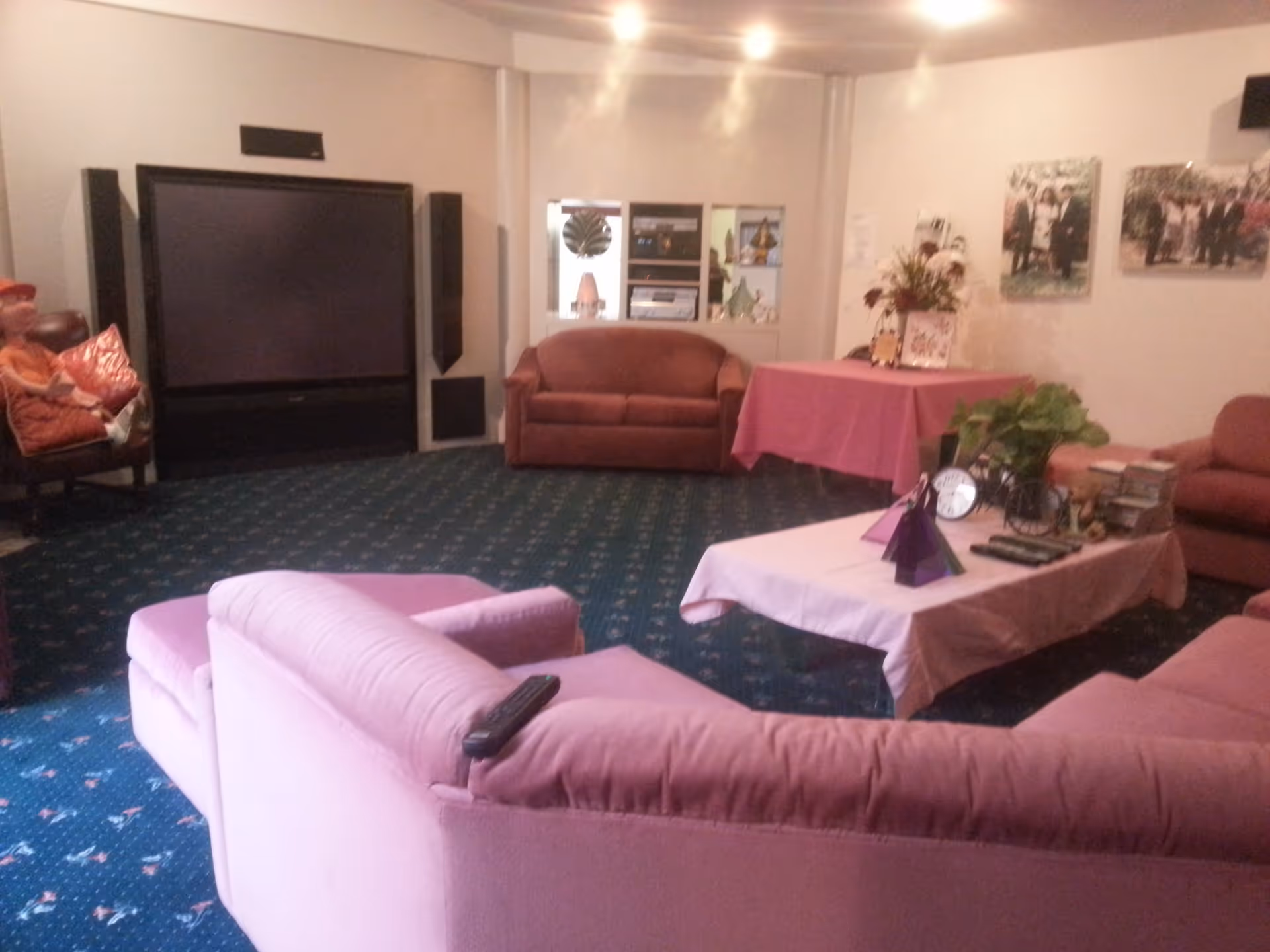 Spacious living room with pink upholstered sofas arranged around a low table, a large TV and speakers against the wall, and framed photos on the right wall.