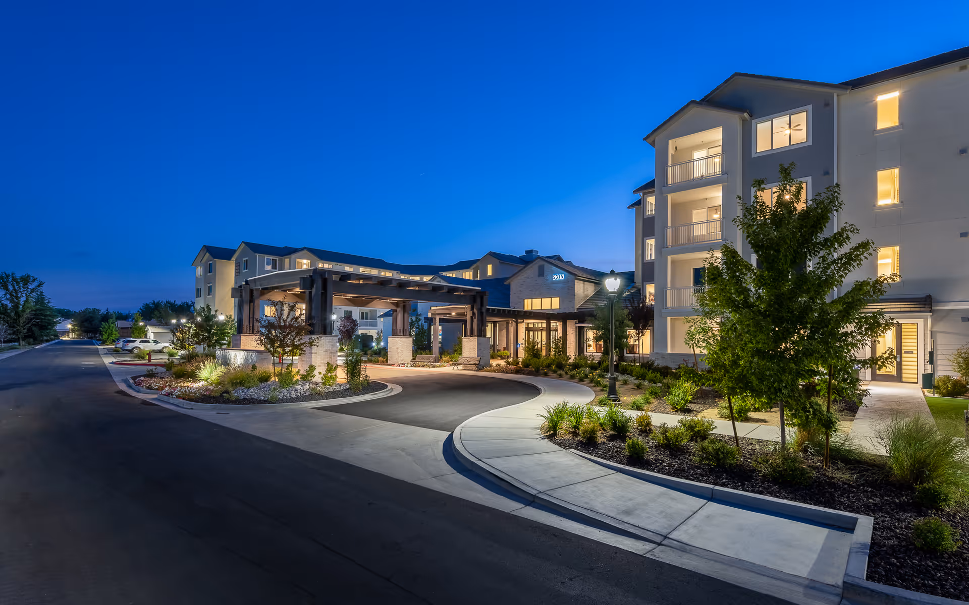 Exterior view of Revel Rancharrah senior living facility at dusk, showing a well-lit entrance with a covered drop-off area, landscaped greenery, and a multi-story building with balconies and large windows.