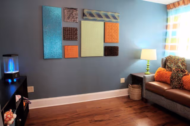 A cozy living room with a gray couch adorned with orange and brown pillows and a multicolored throw blanket. A green lamp sits on a dark wooden side table next to the couch. The wall behind is painted blue and decorated with a series of colorful textured rectangular panels. The floor is wooden, and a black shelving unit with a small illuminated water feature is visible on the left side.
