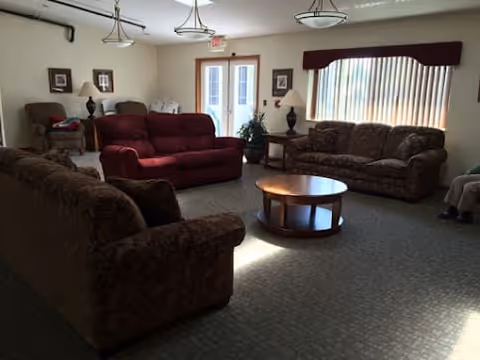 A cozy living room with multiple upholstered sofas and armchairs arranged around a round wooden coffee table. The room has carpeted flooring, large windows with vertical blinds, and ceiling light fixtures. There are framed pictures on the walls and table lamps on side tables.