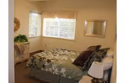 A bedroom with a bed covered in a green floral bedspread and several pillows. There is a window with blinds and a valance, a small dresser with a plant on it, a mirror on the wall, and a bedside table with a lamp.