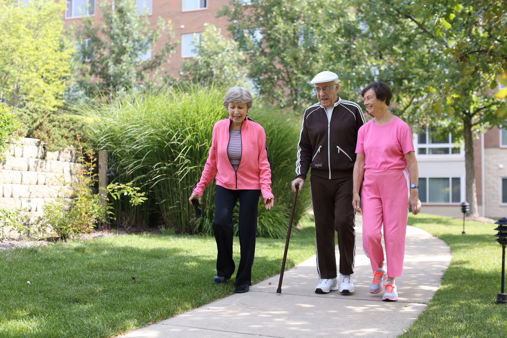Three elderly people walking together on a paved path in a garden area outside a building. Two women are wearing pink outfits, and a man in a brown tracksuit with a white cap is using a cane. The background shows green bushes, trees, and part of a brick building.