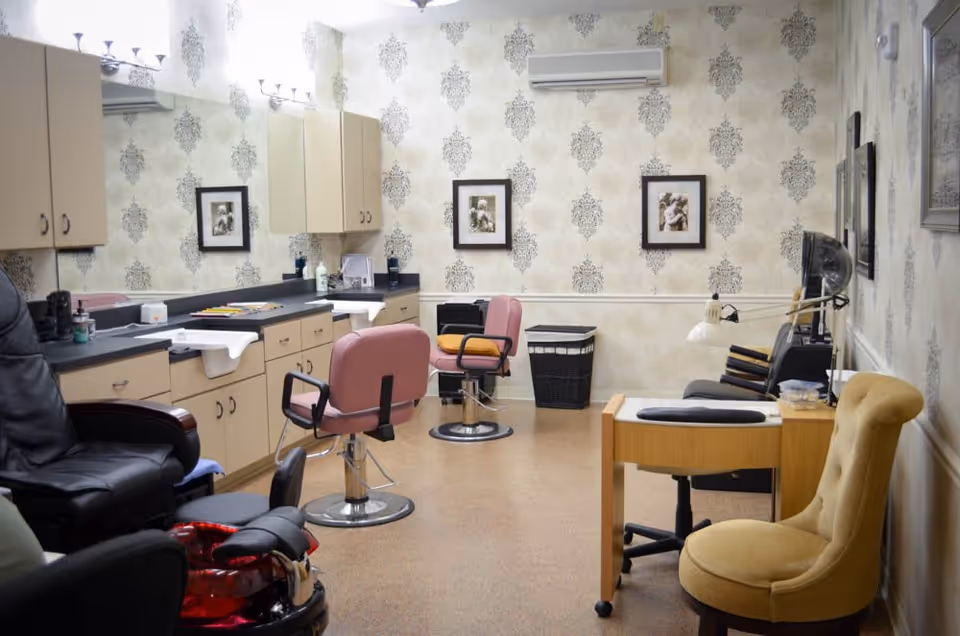 Interior view of a salon or beauty care room with two pink salon chairs, a black pedicure chair with a red foot bath, a manicure table with a yellow chair, and cabinets with sinks along one wall. The walls have patterned wallpaper and framed pictures, and there is an air conditioning unit mounted on the wall.