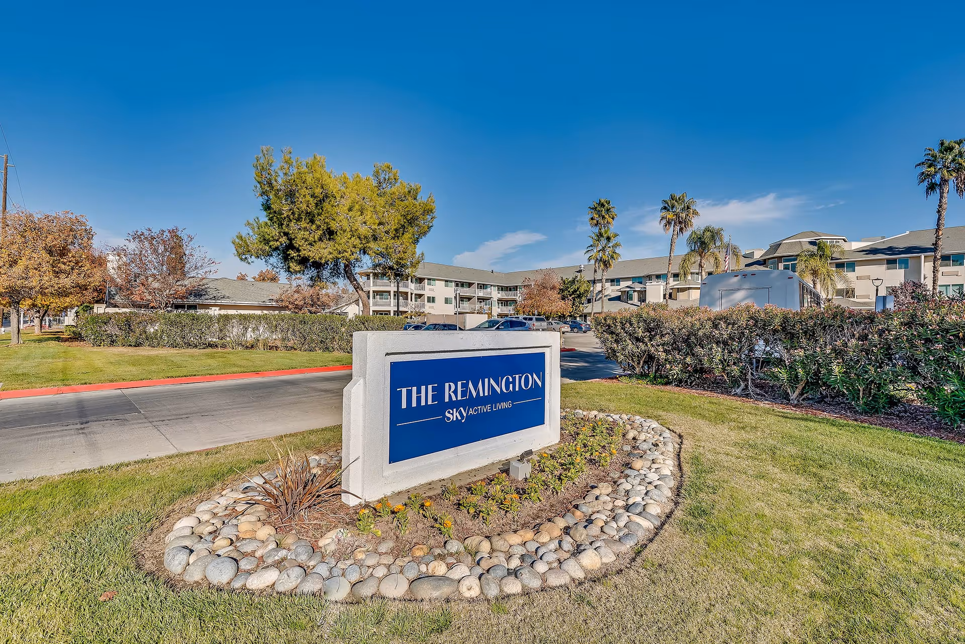 A landscaped entrance sign reading "THE REMINGTON" beside a driveway, with the facility building and palm trees in the background under a clear blue sky.