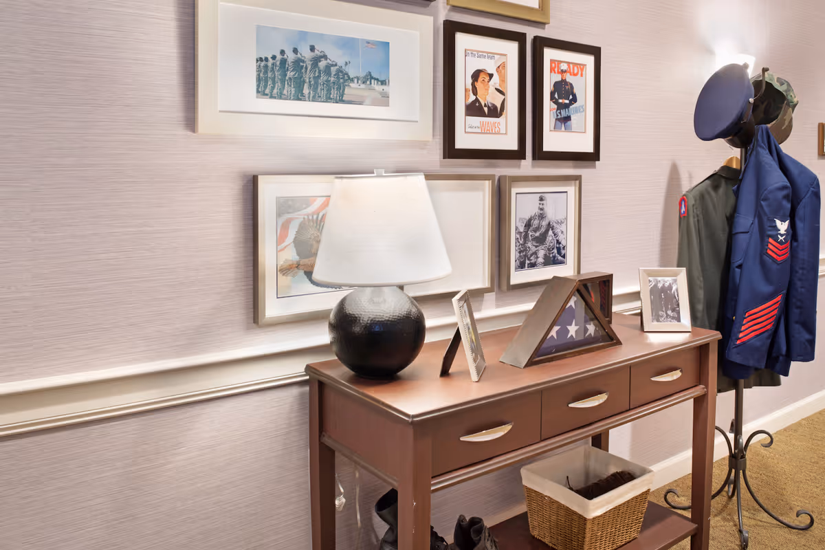 A wooden console table with three drawers and a lower shelf holding a basket and shoes. On top of the table is a black round lamp with a white shade, framed photographs, and a folded American flag in a display case. Above the table, several framed military-themed pictures hang on a light-colored wall. To the right, a coat rack holds military uniforms and hats.