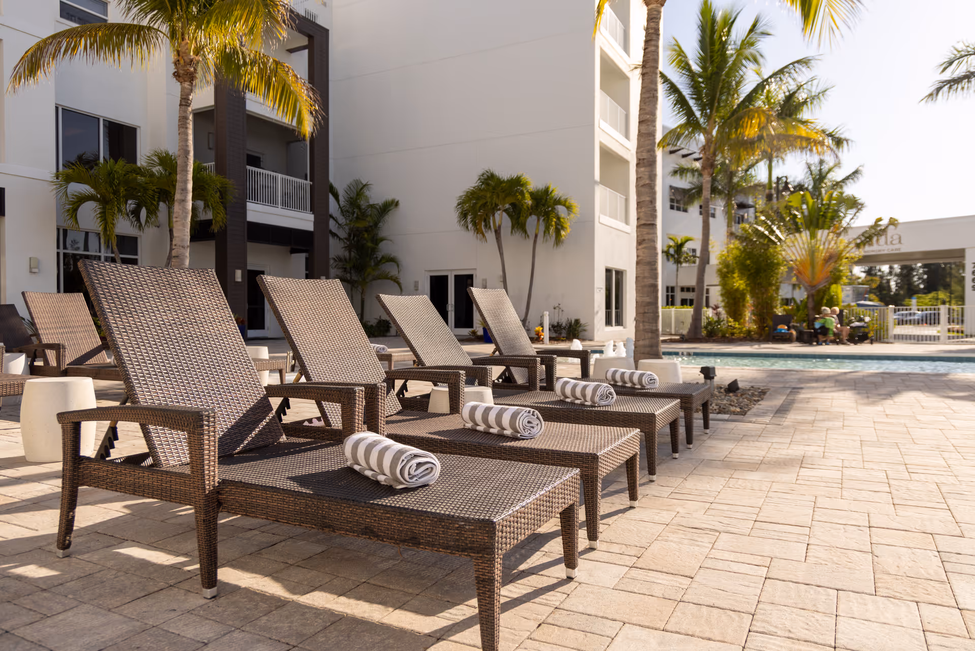 Outdoor pool area at Amavida at Lakes Park featuring several brown wicker lounge chairs with rolled striped towels on them, palm trees, and a multi-story building in the background under a clear sky.