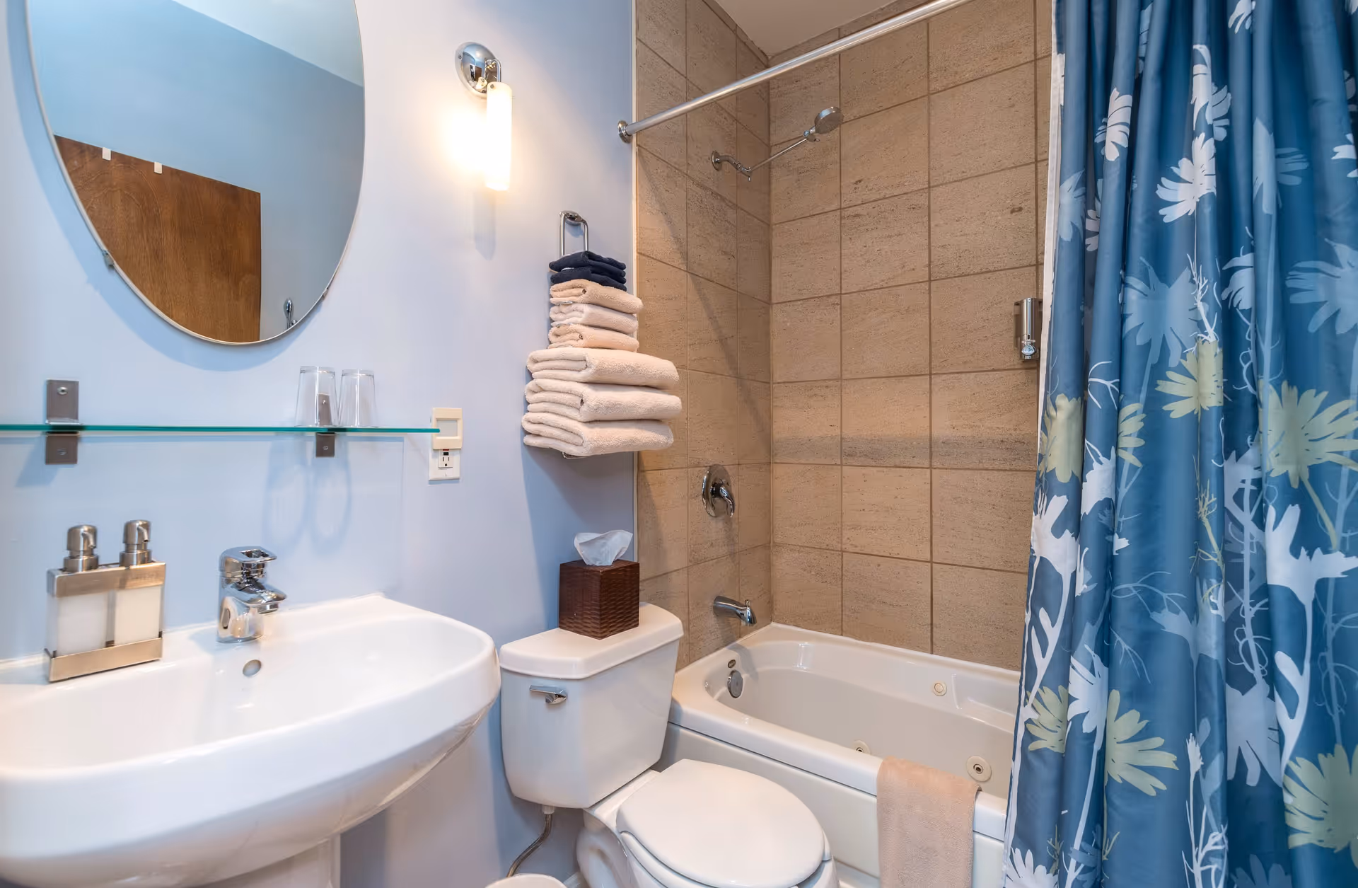 Bathroom with a pedestal sink, toilet, stacked towels, and a bathtub/shower with tiled surround and a blue floral shower curtain.
