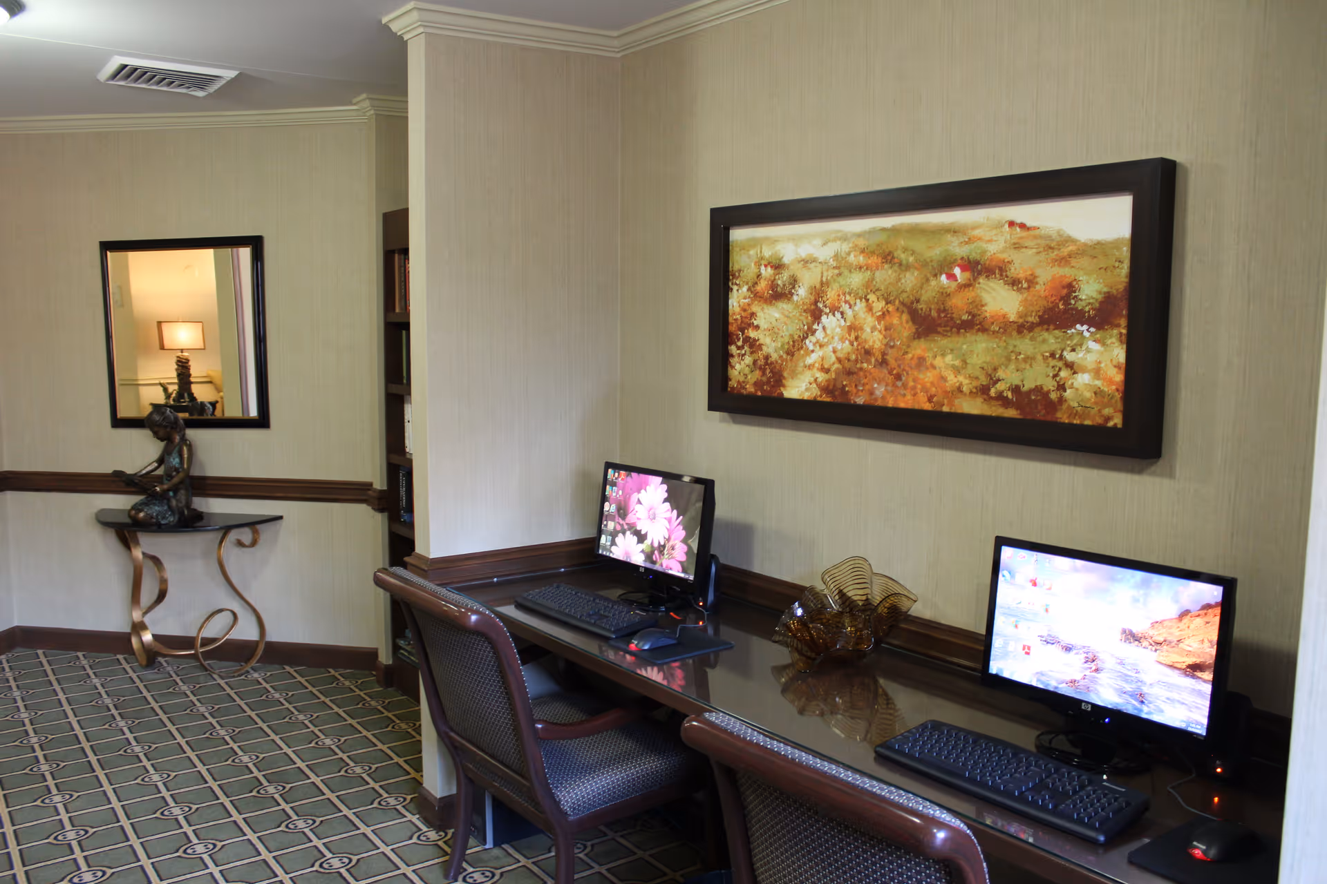 A small computer workstation area in a senior living facility with two desktop computers on a dark wooden desk. Two chairs are placed in front of the computers. Above the desk hangs a framed landscape painting with autumn colors. To the left, there is a decorative table with a sculpture and a mirror on the wall. The room has beige walls and patterned carpet flooring.