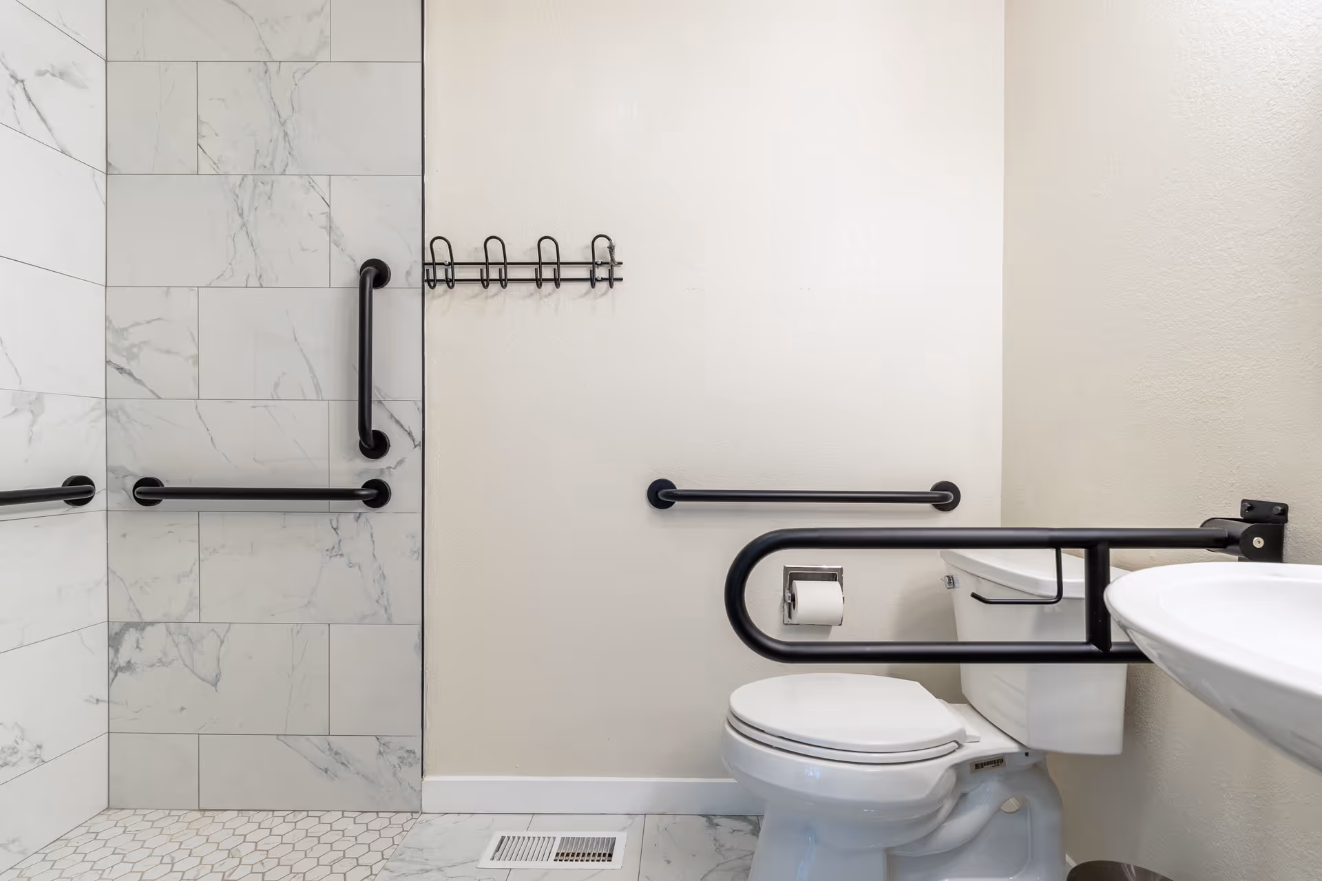Accessible bathroom showing a toilet, wall-mounted sink, multiple grab bars, and a tiled shower area.