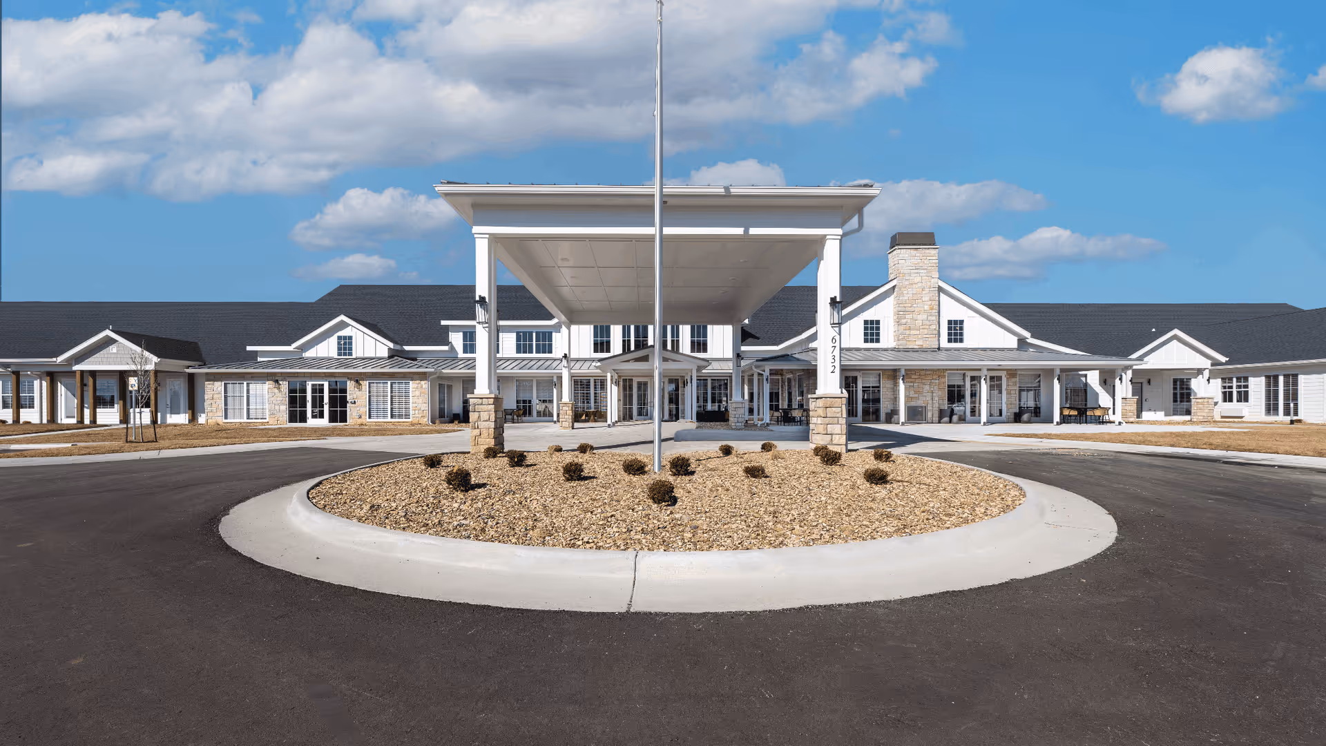 Front exterior view of Cedarhurst Senior Living of Topeka, showing a large building with a covered entrance, stone and white siding, a circular driveway with landscaping in the center, under a partly cloudy blue sky.