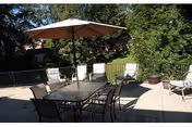 Sunlit outdoor patio with a glass-top dining table, several chairs and lounge chairs, and a large umbrella surrounded by greenery.