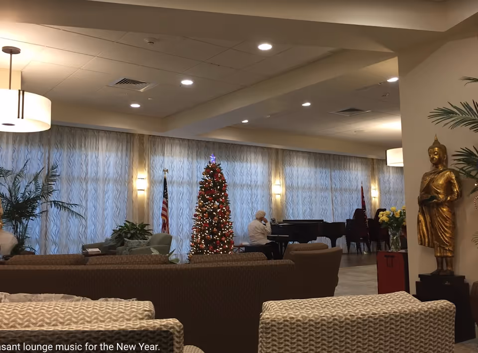 A senior living lounge with seating, a decorated Christmas tree, a person at a grand piano, plants, and a golden statue.