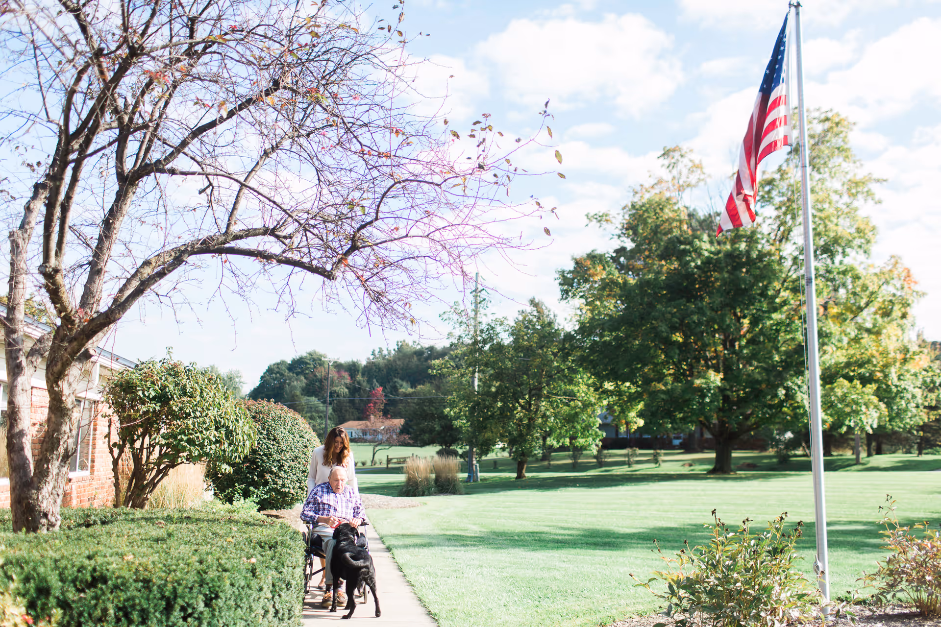A caregiver pushes an older adult in a wheelchair along a sidewalk outside a senior living facility with an American flag and trees on the lawn.