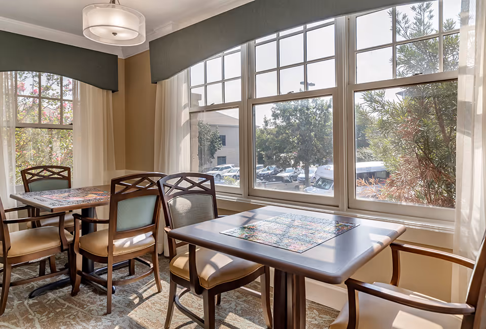 A bright dining area with two wooden tables and several cushioned chairs near large windows that let in natural light. Outside the windows, trees and parked cars are visible. The room has light-colored walls and carpeted flooring.