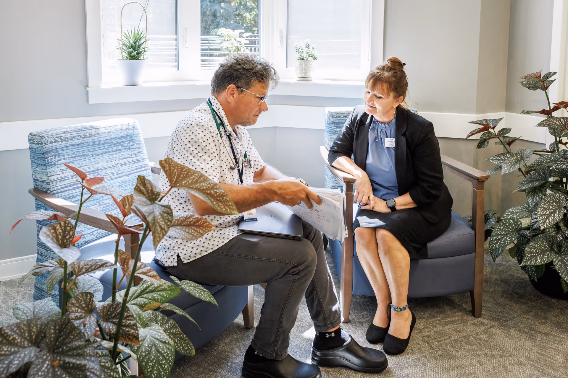 A man and a woman sitting in a well-lit room with large windows and plants. The man, wearing glasses and a patterned shirt with a stethoscope around his neck, is showing papers to the woman, who is dressed in a black blazer and skirt. They are seated on blue cushioned chairs and appear to be engaged in a discussion.
