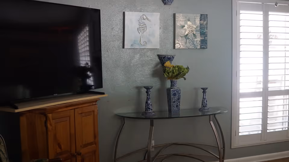 A living room corner featuring a large flat-screen TV on a wooden cabinet, a glass console table with three blue and white decorative vases and a flower arrangement, two framed wall art pieces depicting a seahorse and a starfish, and a window with white plantation shutters.