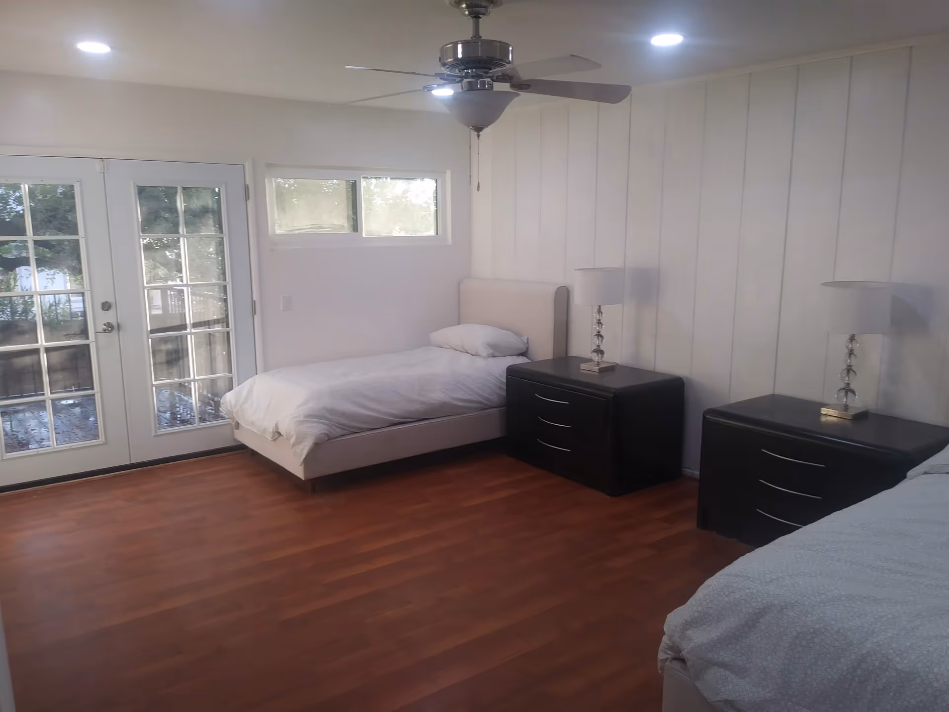 A bedroom with two single beds, each with white bedding and a pillow. Between the beds are two black nightstands, each with a modern lamp. The room has wooden flooring, white paneled walls, a ceiling fan with lights, and a set of glass double doors leading outside. There is also a small horizontal window above one of the beds.