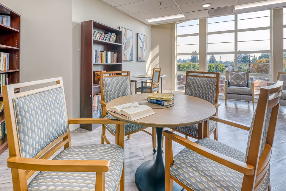 A bright and airy common area in a senior living facility featuring a round wooden table with four patterned cushioned chairs. On the table are a few books and an open book with reading glasses. In the background, there are bookshelves filled with books, additional seating with armchairs, large windows letting in natural light, and a view of trees and rooftops outside.