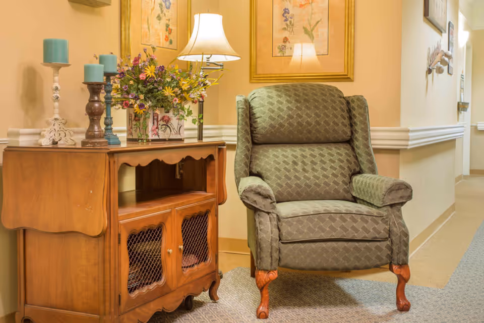 A cozy corner in a senior living facility hallway featuring a patterned green armchair with wooden legs next to a wooden side table. The table holds a lamp, three blue candles on candle holders, and a floral arrangement. The walls are decorated with framed botanical prints.