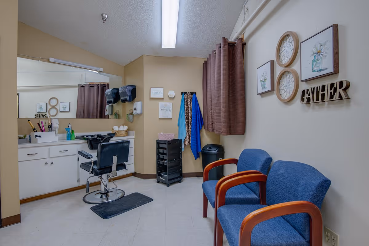 Interior view of a small salon or grooming area with a black salon chair in front of a counter with a sink, hair care products, and a large mirror. On the right side, there are two blue chairs with wooden armrests, brown curtains on the window, wall decorations including framed pictures and the word 'GATHER'. A black trash bin and a rolling cart with supplies are also visible.