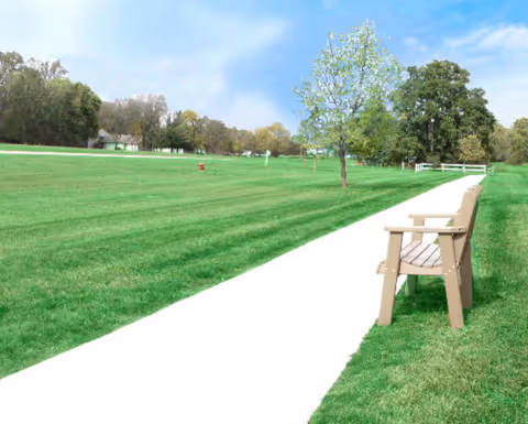 A paved walkway runs through a large green grassy area with a wooden bench on the right side. There are several trees scattered around the lawn and a few buildings visible in the distance under a partly cloudy sky.