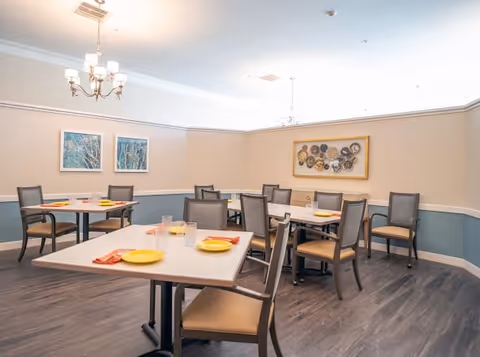 A dining room with several square tables set with yellow plates, orange napkins, and clear drinking glasses. The room has beige walls with a blue lower section and white trim, decorated with framed artwork and a chandelier hanging from the ceiling. The floor is dark wood.