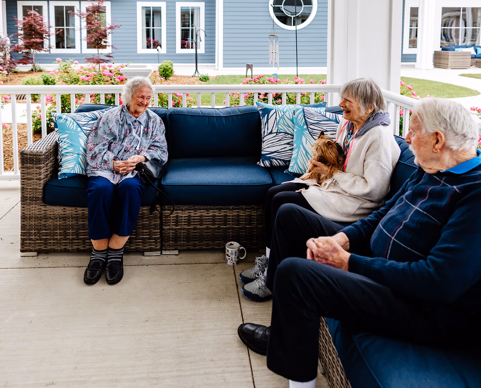 Three older adults sit on wicker patio sofas under a covered porch, two women chatting (one holding a small dog) and a man seated at the right with a garden and building visible behind them.