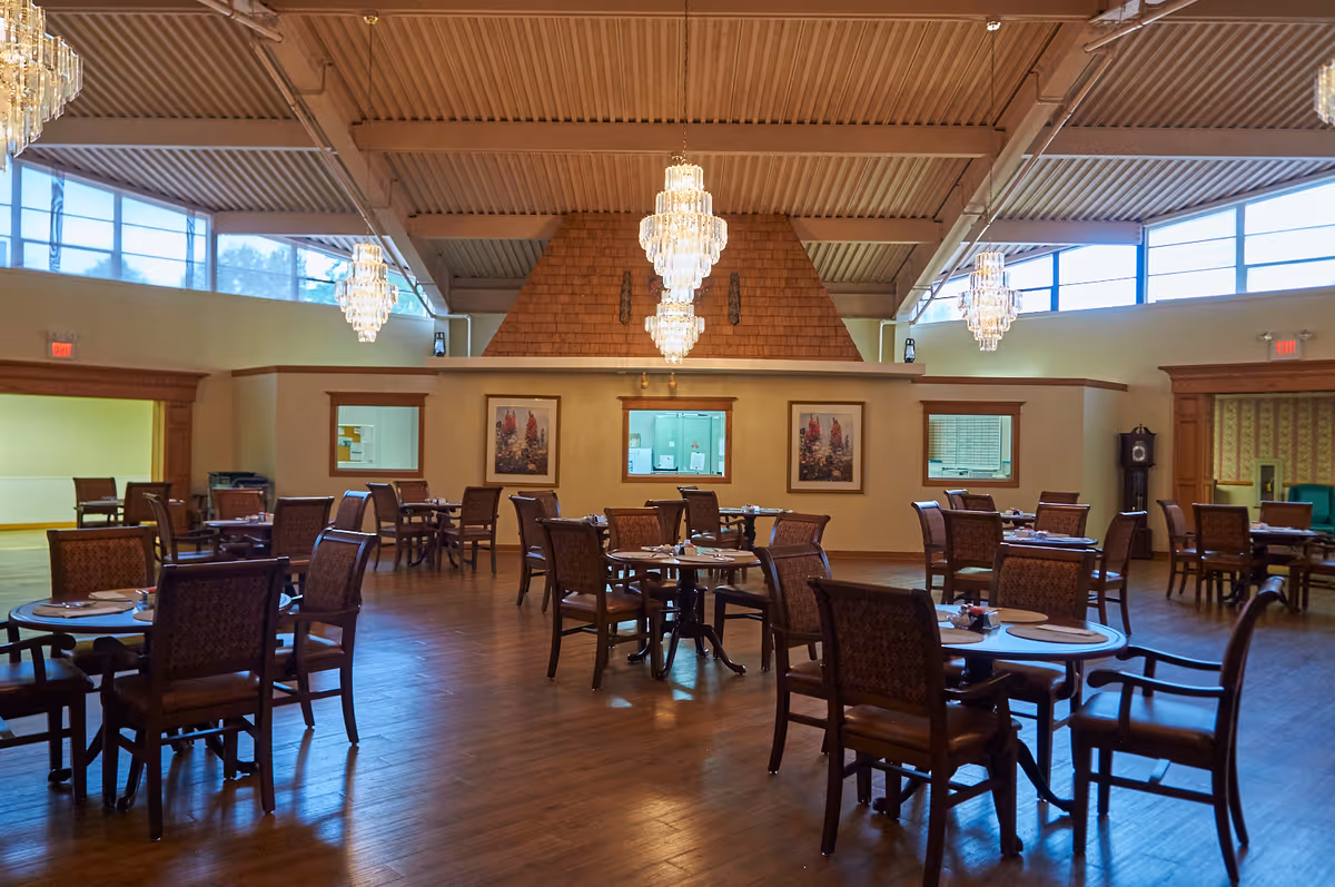 A spacious dining room with multiple round wooden tables and cushioned chairs arranged neatly. The room features large windows near the ceiling allowing natural light, elegant chandeliers hanging from the ceiling, wooden flooring, and framed artwork on the walls. There is a clock in the corner and an open doorway leading to another room.