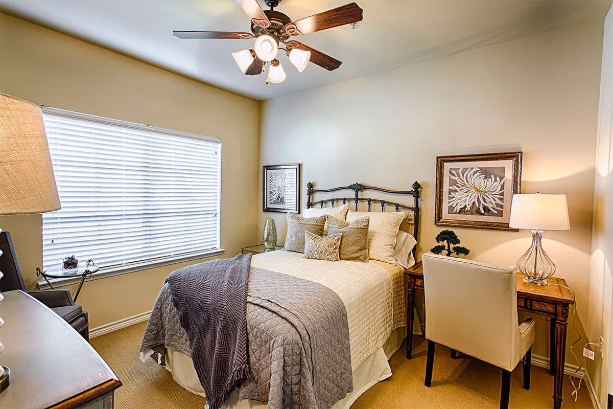 A cozy bedroom with a neatly made bed featuring multiple pillows and a gray quilted blanket. The room has beige walls, a ceiling fan with lights, a window with white blinds, a wooden desk with a lamp and a small plant, and a white upholstered chair. Two framed floral artworks hang on the walls.