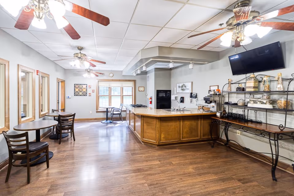 Interior view of a communal dining and kitchen area in a senior living facility. The room features wooden floors, several small round tables with chairs, ceiling fans with lights, a kitchen island with a granite countertop, a wall-mounted TV, and a shelving unit with jars and decorative items. Large windows allow natural light into the space.