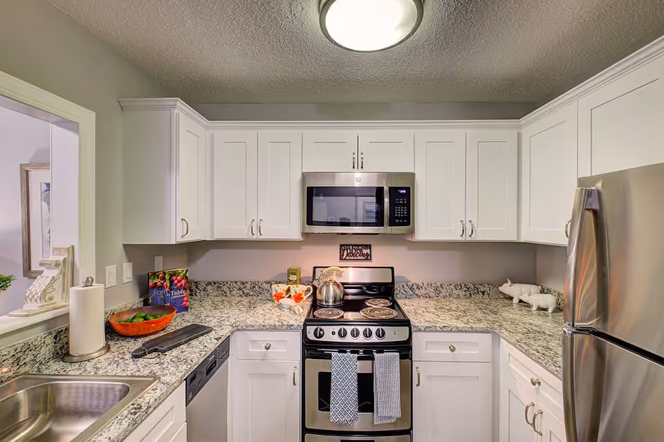 A modern kitchen with white cabinets, granite countertops, a stainless steel refrigerator, a stove with a kettle on it, a microwave above the stove, a dishwasher, a sink, and various kitchen items including a bowl of limes, a paper towel holder, and decorative pieces.