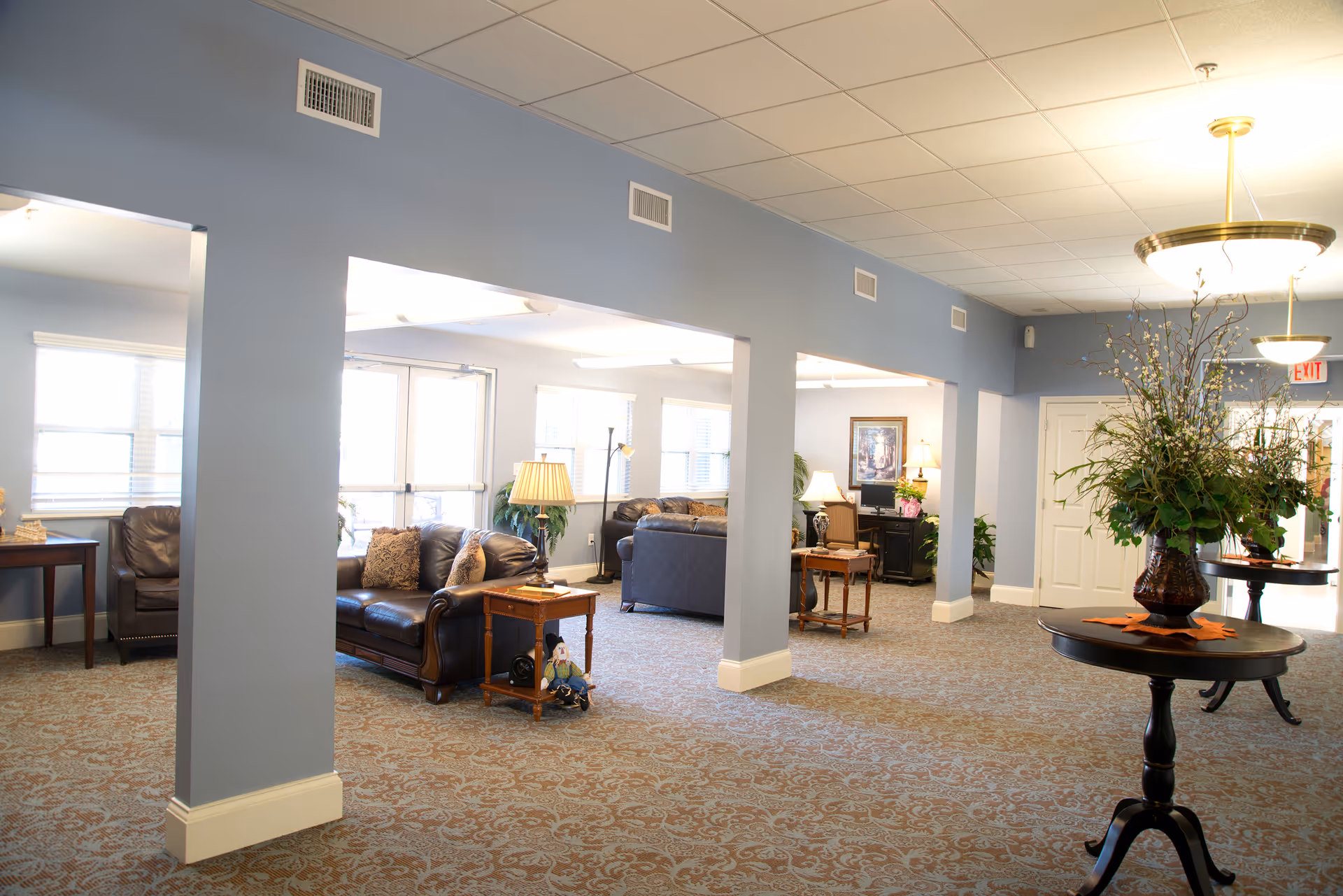 A spacious and well-lit common area in a senior living facility with blue walls and patterned carpet. The room features several leather sofas, wooden side tables with lamps, decorative plants, and framed artwork on the walls. There are large windows letting in natural light and a round table with a large floral arrangement in the foreground.