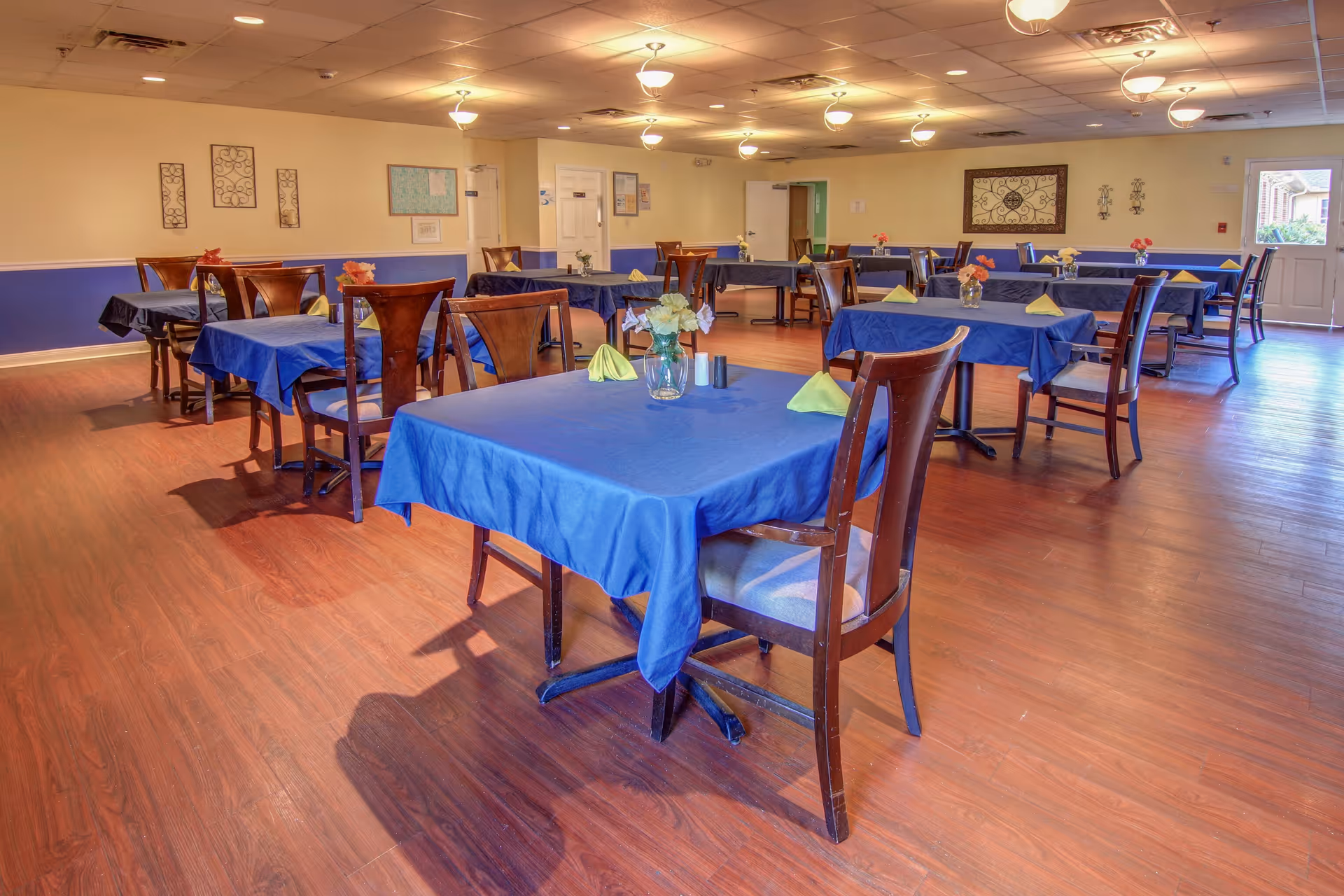 A dining room with multiple tables covered in blue tablecloths, each set with folded yellow napkins and small flower vases. Wooden chairs surround the tables, and the room has wood flooring, beige walls with a blue lower section, and ceiling lights.