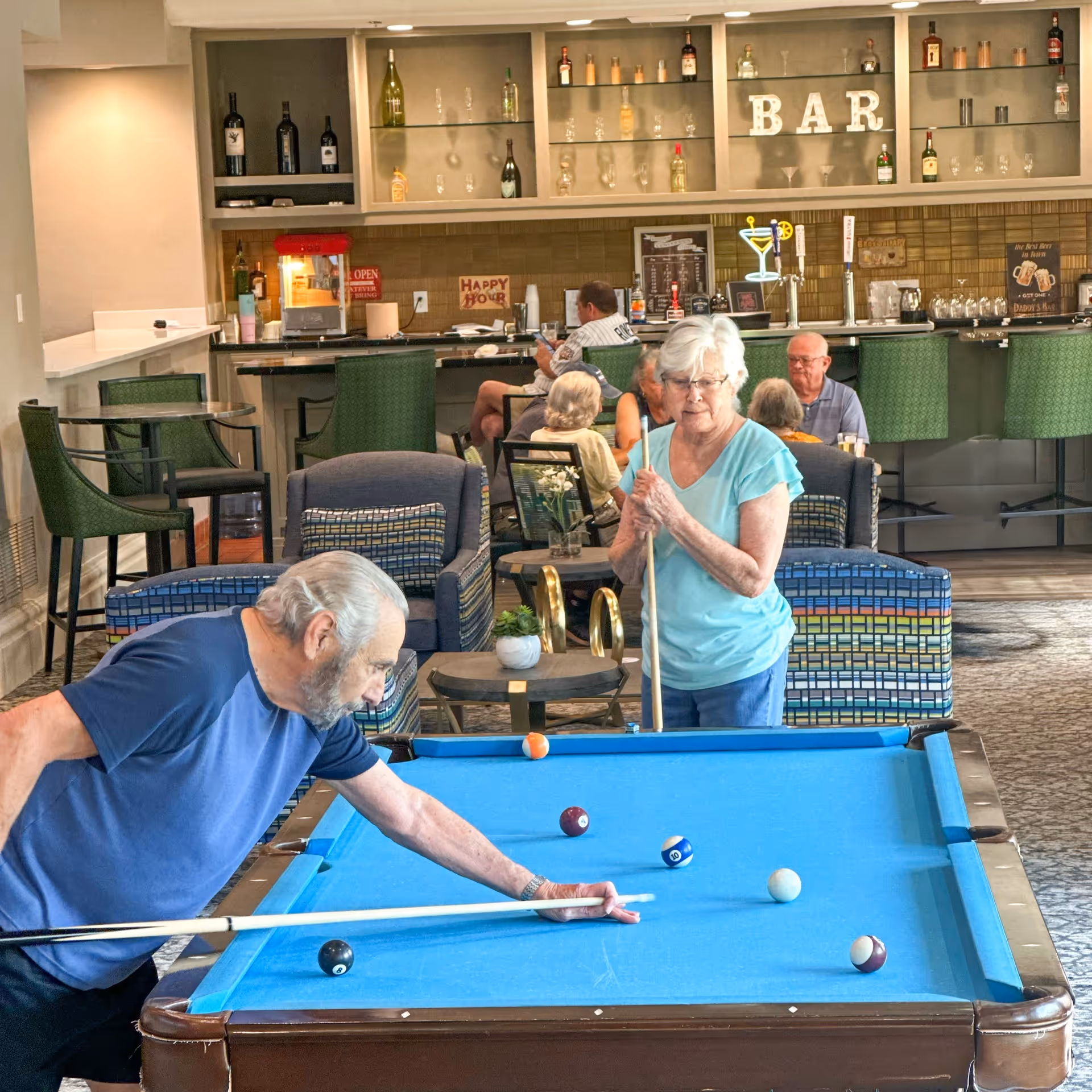Two elderly people playing pool in a senior living facility lounge area with a bar in the background. Several other seniors are seated and socializing near the bar, which is stocked with bottles and glasses. The room has comfortable chairs and a relaxed atmosphere.