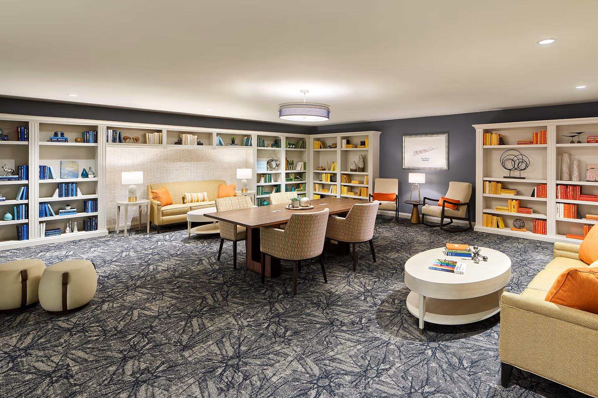 A spacious, well-lit common room with patterned carpet, a large wooden table surrounded by six upholstered chairs, two beige sofas with orange cushions, two beige armchairs with orange pillows, and multiple white bookshelves filled with color-coordinated books and decorative items. The walls are painted gray, and there are several table lamps and a ceiling light fixture providing warm lighting.
