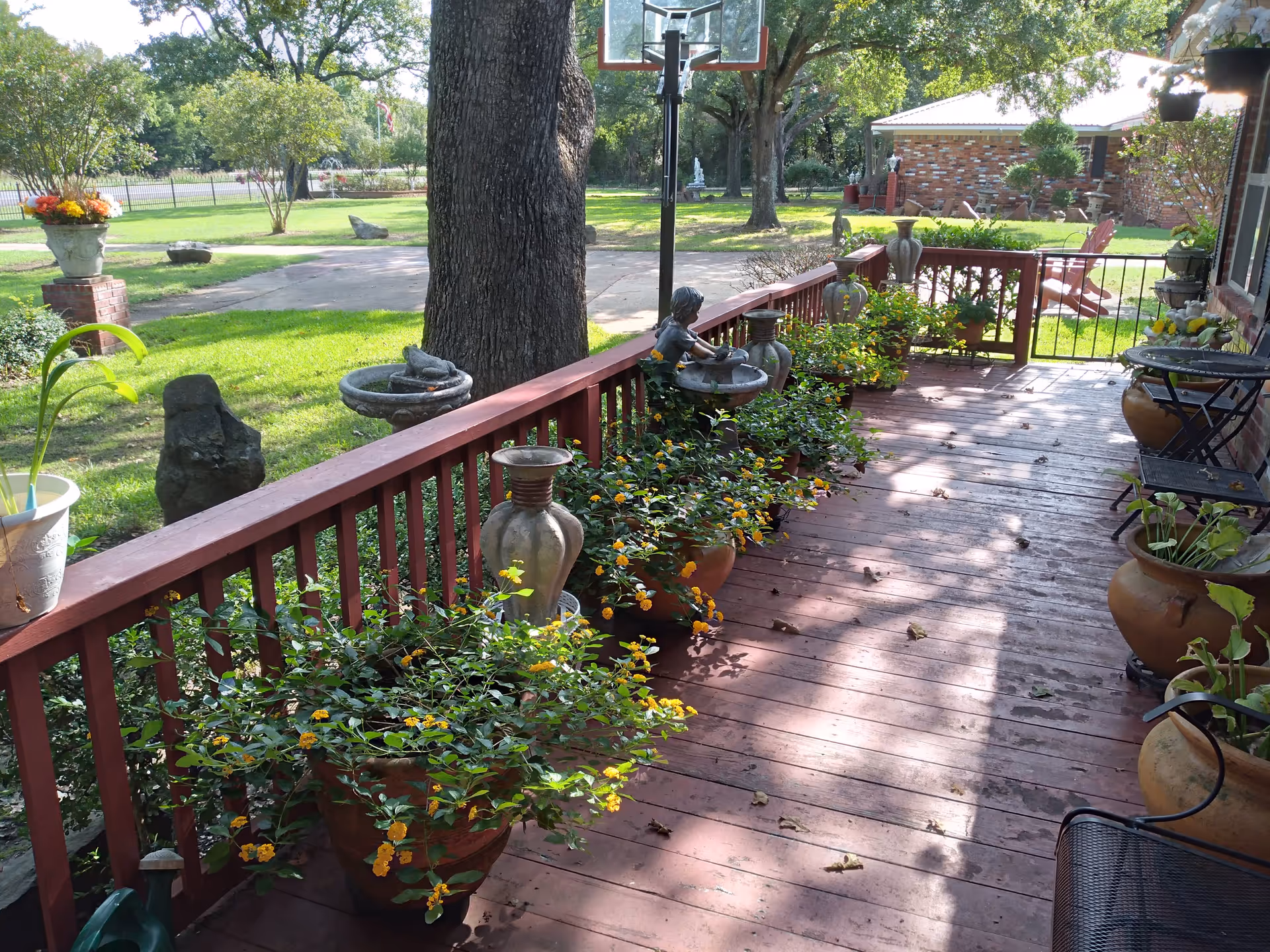 Sunlit wooden front porch lined with potted plants, decorative urns, and seating overlooking a grassy yard and driveway.