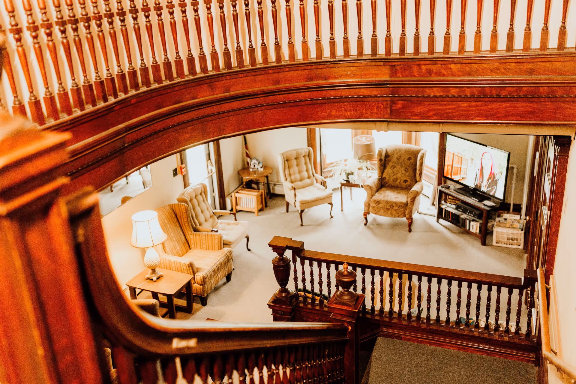 View from the top of a wooden staircase looking down into a cozy living room area with several upholstered armchairs, a side table with a lamp, a TV on a stand, and large windows letting in natural light.