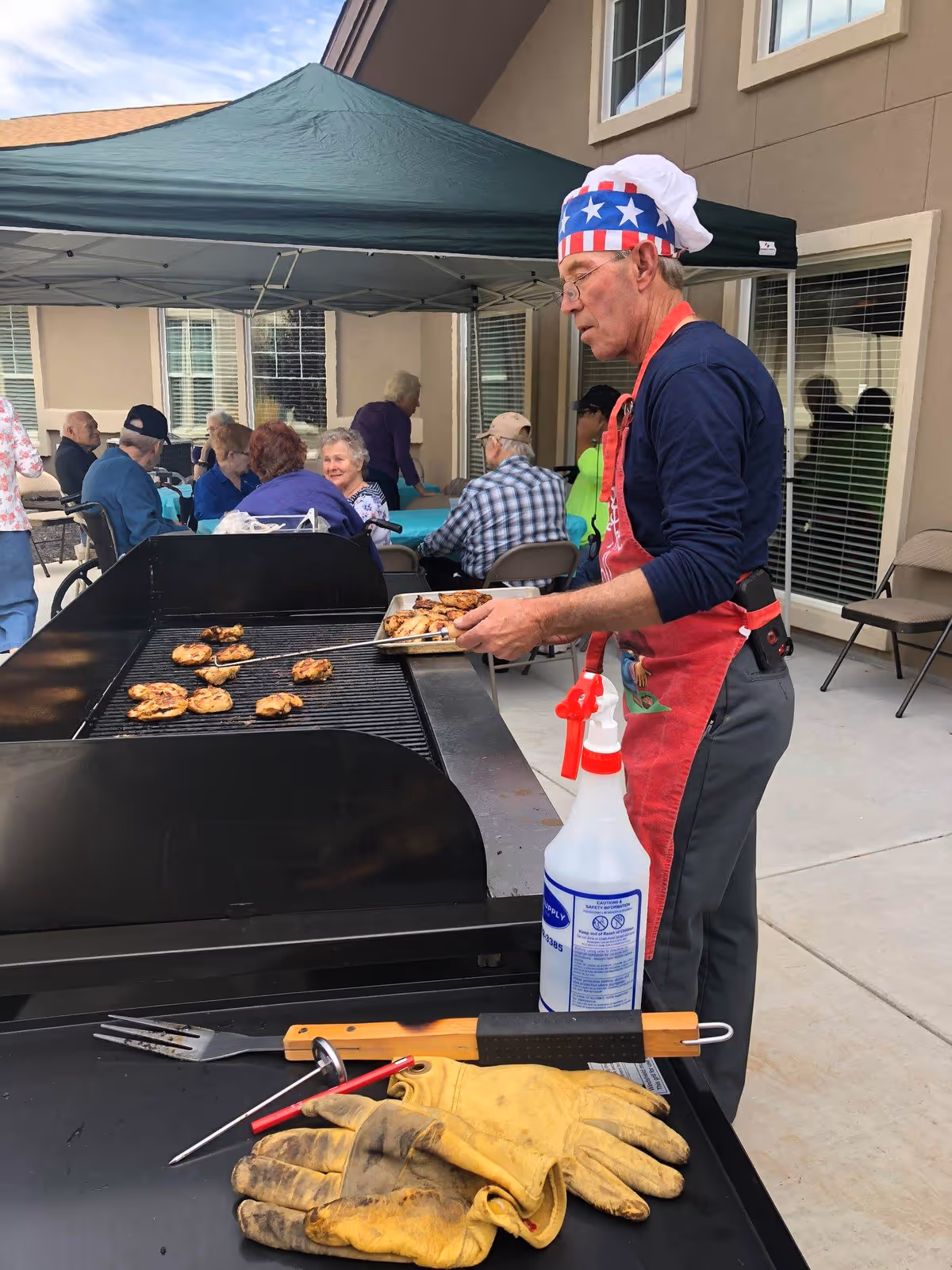 An elderly man wearing a patriotic chef hat and red apron grills chicken on a large outdoor barbecue grill. Several elderly people are seated at tables under a green canopy in the background, enjoying a social gathering outside a building.