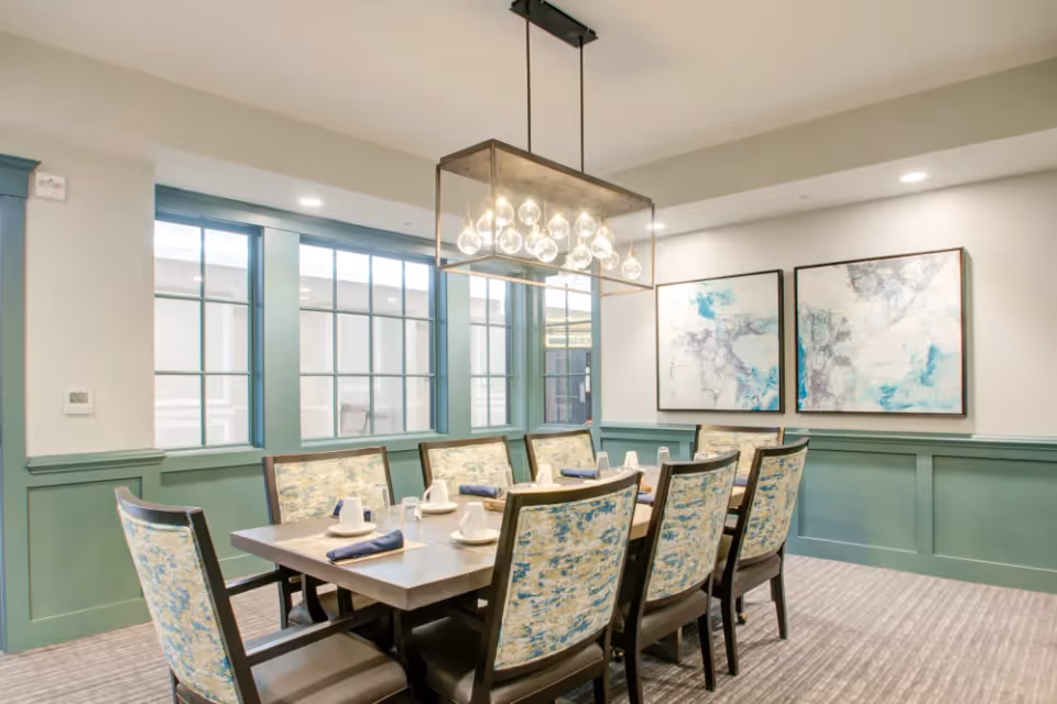 Well-lit dining room with a rectangular table surrounded by upholstered chairs, a modern pendant light, and abstract wall art.